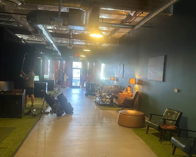 The image shows a spacious hallway with green carpet, multiple seating areas, and individuals engaging in various activities, including golf practice.