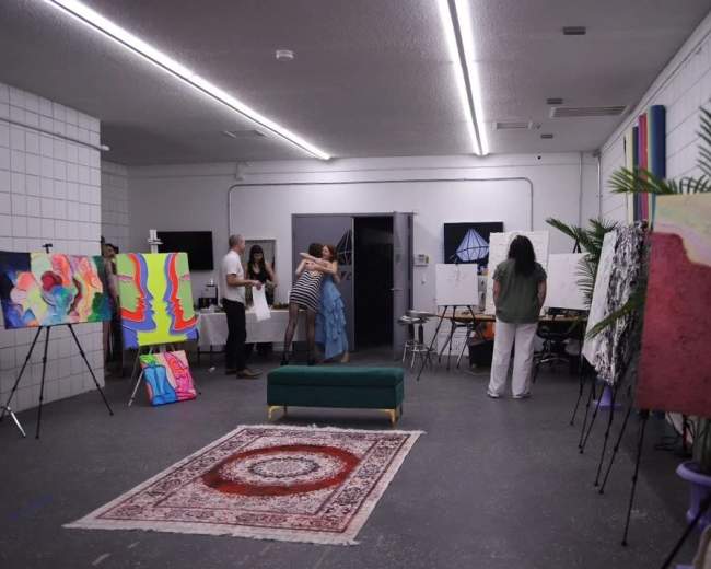 A group of people interacts in an art gallery featuring several colorful paintings displayed on easels and walls, with a patterned rug in the center.