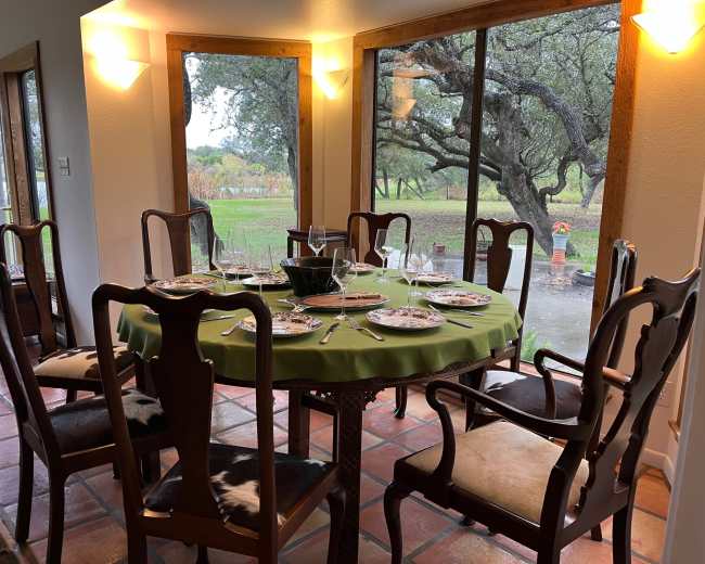 A dining table is set with dishes and glassware in a room featuring large windows that overlook a rainy outdoor scene.
