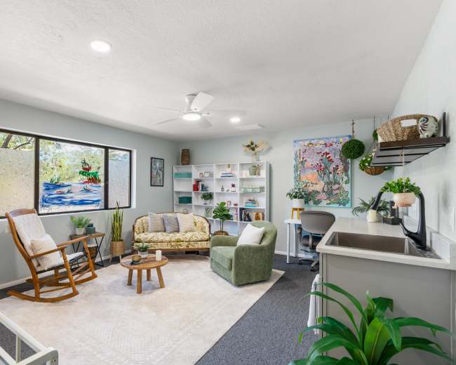 The image shows a modern living room with a green couch, a rocking chair, a small coffee table, and colorful wall art near a large window.