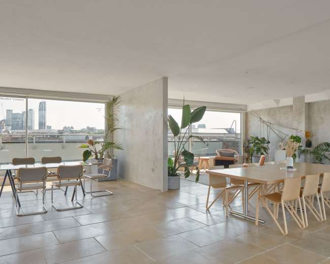 The image shows a spacious indoor area with a large dining table, chairs, and large windows overlooking a cityscape.