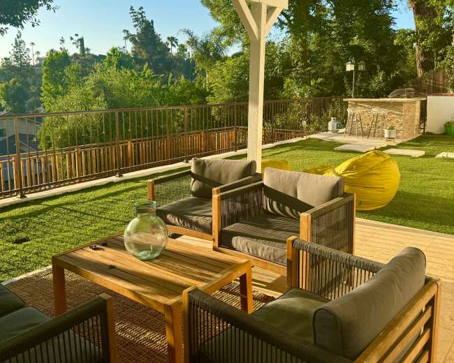 The image shows an outdoor living space with a wooden table and seating arranged under a covered patio, surrounded by greenery and a well-maintained lawn.