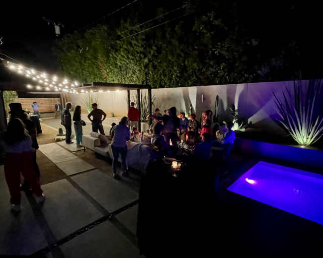 A group of people gathers in a dimly lit backyard, with string lights illuminating the area and a small pool glowing in the corner.