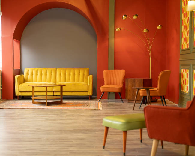 The image shows a modern interior with a yellow couch, orange chairs, and a floor lamp against a backdrop of red and gray walls.