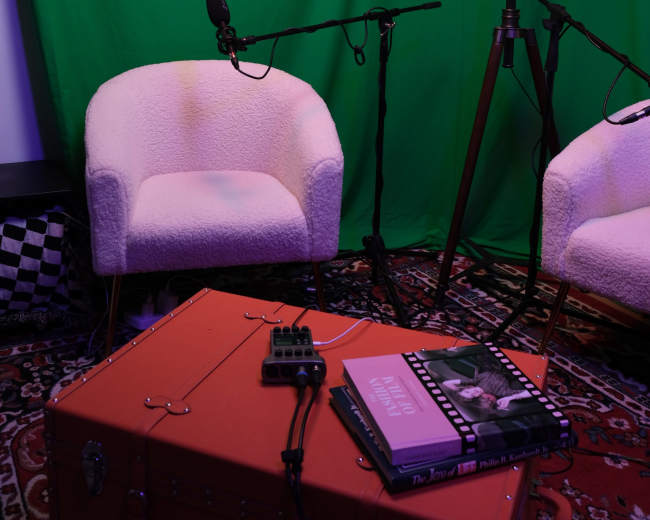 The scene features two white chairs positioned in front of a green screen, with microphones set up nearby and an orange suitcase on the floor holding books and cables.