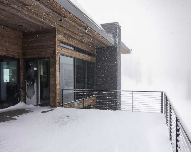 A modern wooden cabin with large windows and a balcony overlooks a snowy landscape shrouded in fog.