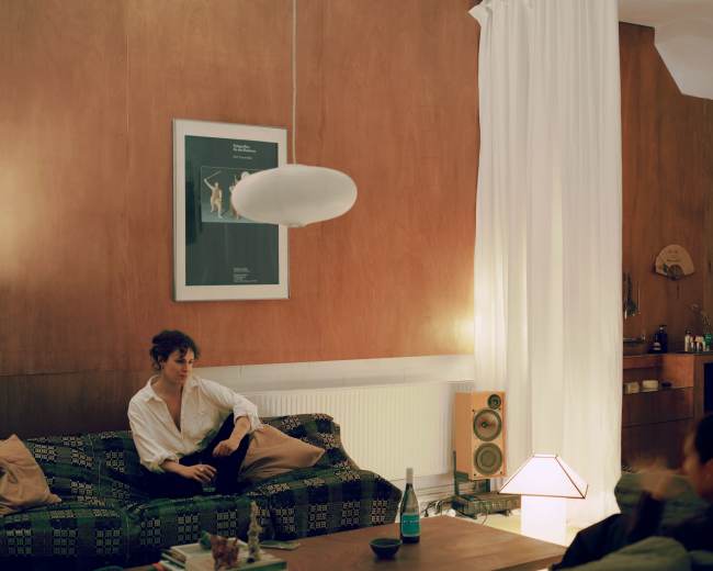 A person sits on a patterned sofa in a warmly lit living room featuring wooden walls, a poster, a floor lamp, and a plant.