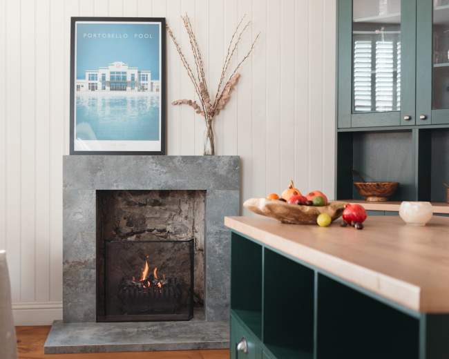 A modern kitchen features a stone fireplace with a small fire, a large framed pool-themed artwork on the wall, and a wooden countertop displaying a bowl of fruit.