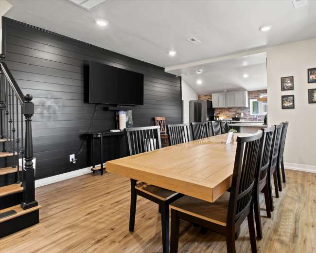 The image shows a dining area with a large wooden table surrounded by black chairs, a television mounted on the wall, and a staircase in the background.