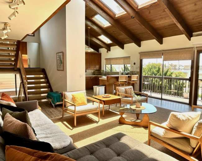 The image shows a spacious living room with vaulted wooden ceilings, large windows, and a staircase leading to an upper level.