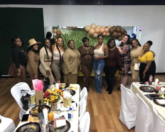 A group of women dressed in similarly colored outfits pose together in front of a decorated backdrop at a festive indoor event.