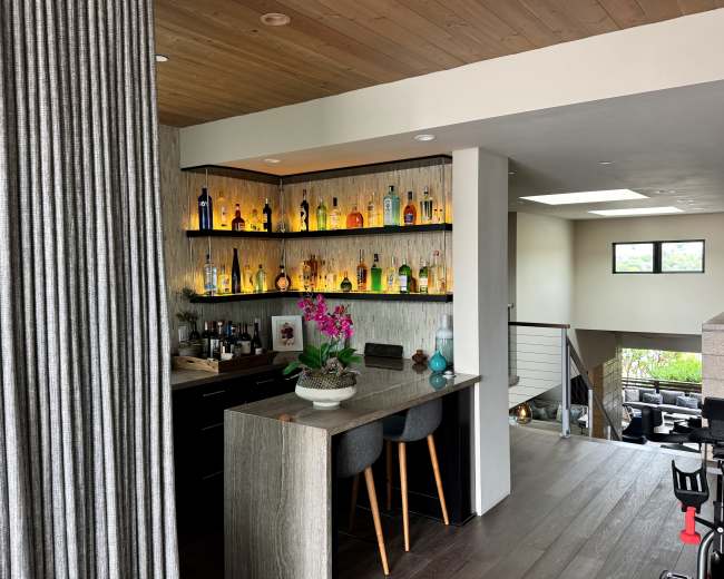 The image shows a modern home interior featuring a sleek bar area with shelves displaying various bottles, complemented by a small decorative plant and seating.