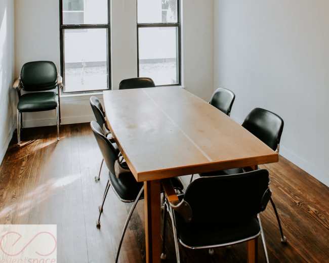 A simple meeting room with a wooden table surrounded by black chairs, featuring two large windows and a ceiling fan.
