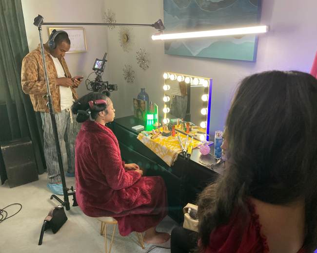 A woman sits at a vanity table with bright lights, while a man adjusts equipment and another woman stands beside her.