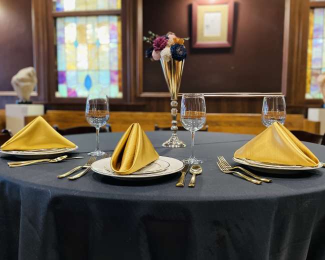 A banquet table set with gold napkins, elegant dishware, glassware, and a floral centerpiece, against a backdrop of stained glass windows.