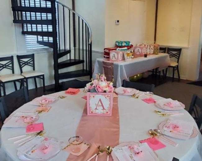 A round table set with white and pink decorations is placed in a room featuring a spiral staircase and a buffet table in the background.