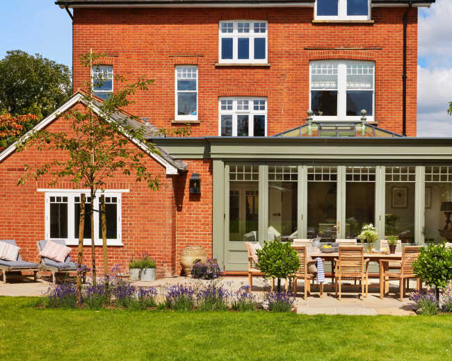 A brick house with large windows features a glass conservatory and a patio with wooden dining furniture surrounded by green grass and flower beds.