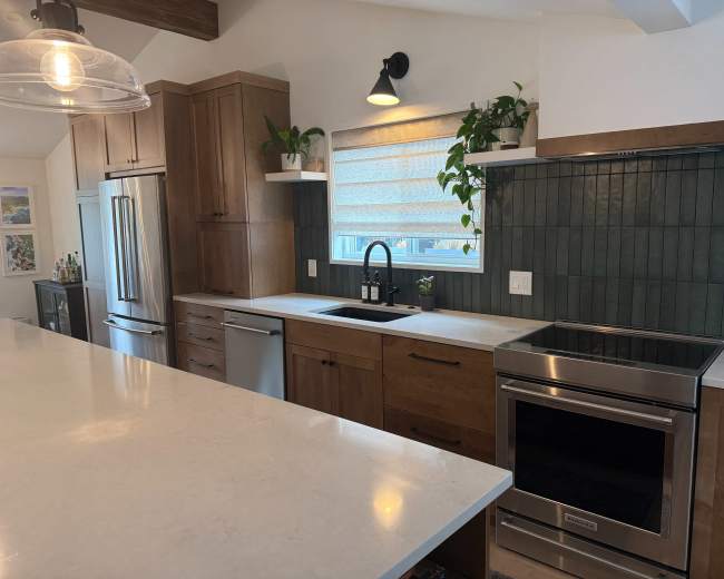 The image shows a modern kitchen with wooden cabinetry, a stainless steel refrigerator, and a countertop made of light-colored stone, featuring an integrated sink and a stove.
