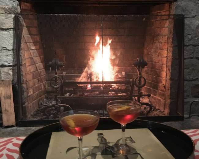 Two cocktail glasses sit on a tray in front of a roaring fireplace, with orange garnishes and a decorative paper underneath.