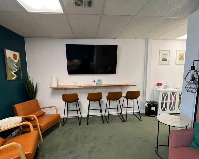 The image depicts a modern lounge area featuring two orange armchairs, a pink side table, a long counter with four bar stools, a wall-mounted television, and a green carpet.