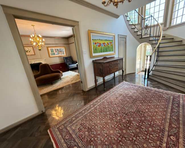 The image shows an entryway with a decorative rug, a wooden console table, and a staircase leading to an upper level, with a glimpse of a living area visible through an archway.