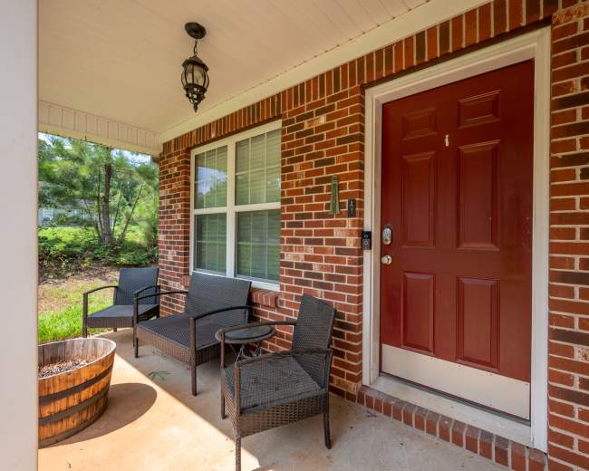 A brick front porch with a red door, two chairs, and a small table beside a wooden barrel.