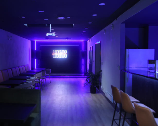 The image shows a modern lounge with purple neon lighting, featuring rows of tables and chairs along the walls.