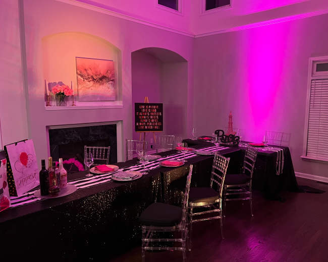 A dining area is set with a black and silver tablecloth, adorned with decorative items, and illuminated by pink lighting.