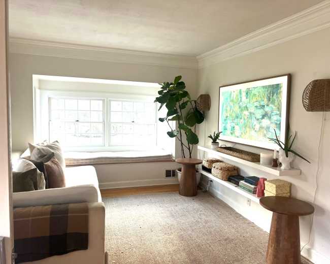 A bright corner of a living room features a white sofa, a large window seat, and a painting above a white shelf with decorative items.