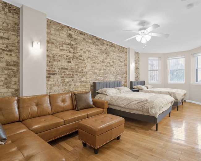 The image shows a spacious bedroom featuring two large beds, a brown leather sofa, and a wooden TV stand against a wall with exposed brick.