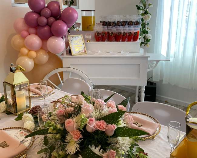 A decorated table with a floral centerpiece and pink-themed place settings is set up beside a balloon arch and a beverage station.