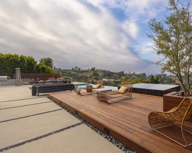 The image shows a wooden deck on a hillside with modern outdoor furniture and a view of a landscape under a cloudy sky.