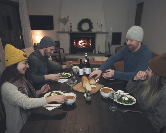 A group of four people shares a meal at a wooden dining table near a fireplace.