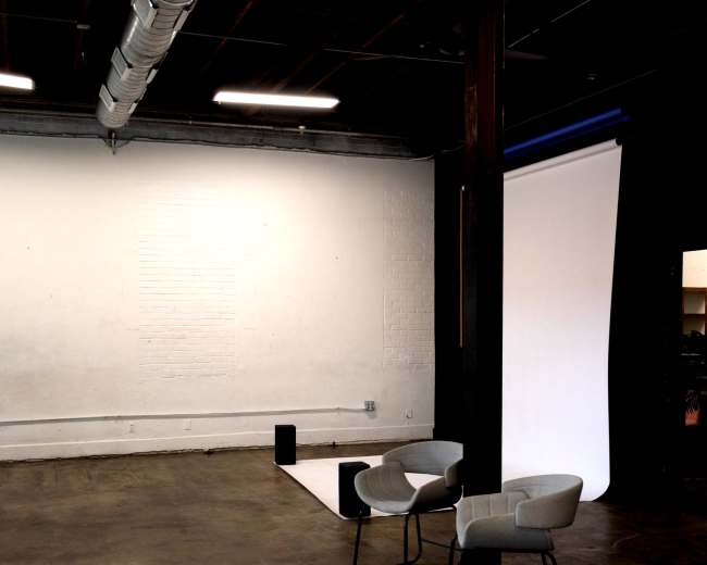 The image depicts an empty studio space featuring a pair of modern chairs, a white backdrop, and industrial-style elements.