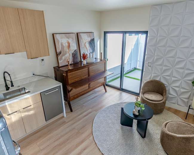 The image shows a modern living space featuring a kitchen area with cabinets, a dining table, and two brown armchairs facing a round table, with a sliding glass door leading to an outdoor patio.