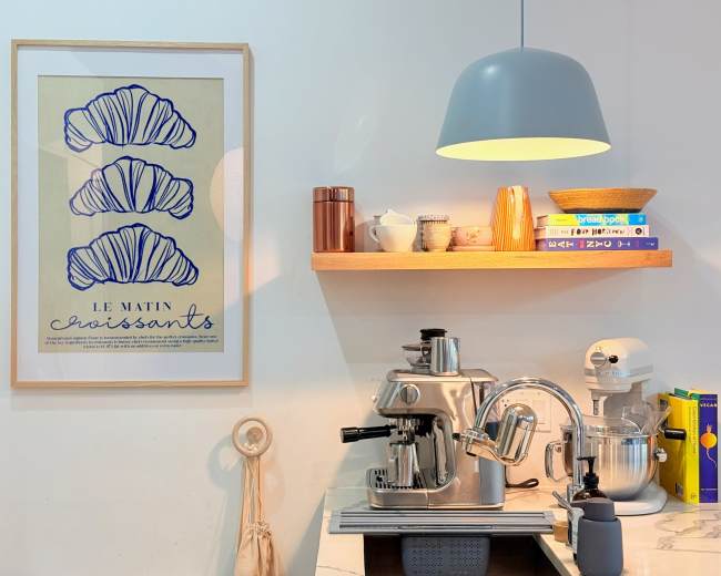 The image shows a modern kitchen corner with a coffee machine, a stand mixer, and a shelf displaying books and decorative items, all beneath a pendant light.