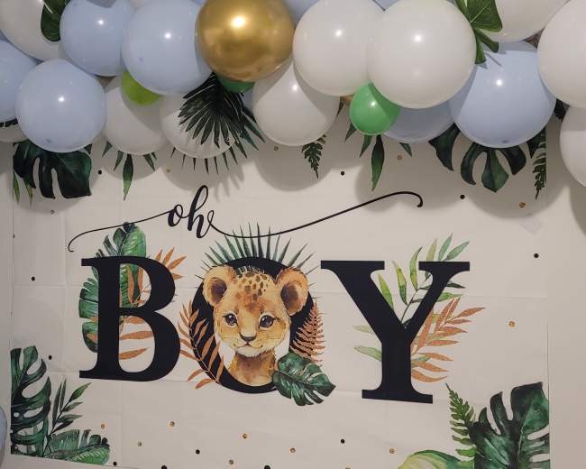 A decorated wall with a banner reading "oh BOY" featuring a lion cub and surrounded by a variety of balloons in green, blue, white, and gold.