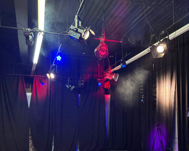 The image shows a stage area with black curtains, lighting equipment, and wooden flooring.