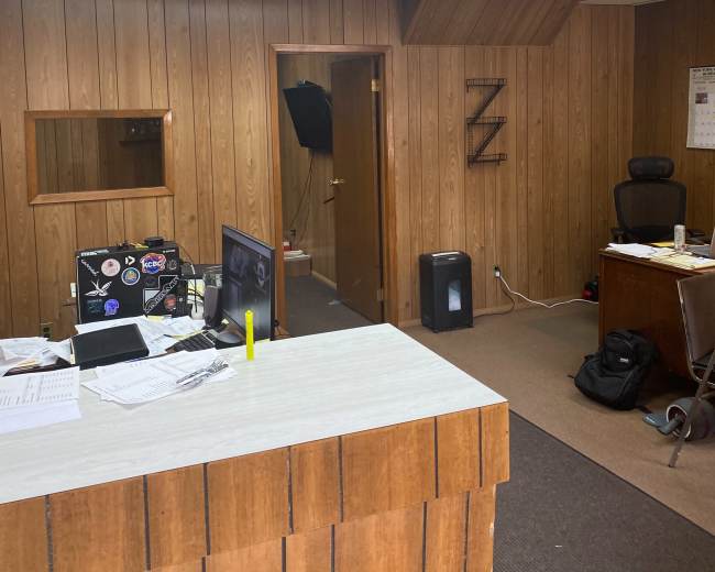 The image shows an office interior with wooden paneling, a reception desk, a chair, a doorway leading to another room, and scattered papers on the surfaces.