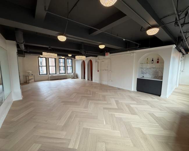 The image shows a bright, modern interior space with herringbone wooden flooring, large windows, and elegant light fixtures.