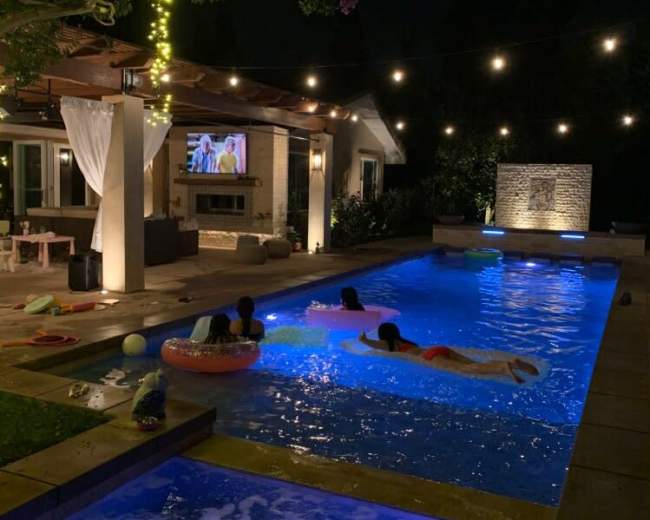 Four people float in a brightly lit pool at night while a television screen displays an image under string lights and surrounded by greenery.