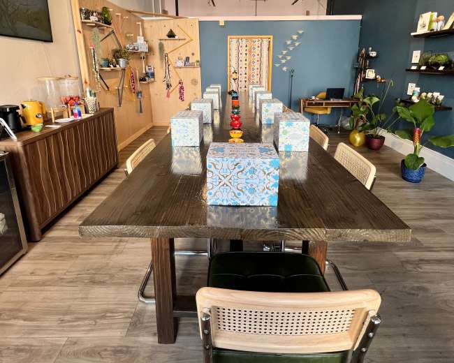 A long wooden table with eight seats is set in a bright room decorated with plants, art, and colorful boxes.