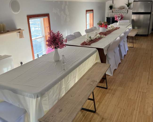 A long table is set up in a room with white walls, featuring pink flower arrangements and wooden benches along the sides.