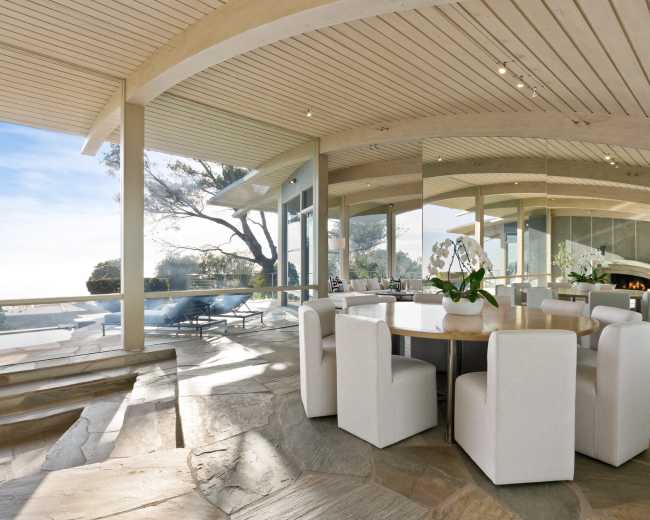 A dining area with a circular table and white chairs is situated next to large glass windows that offer a view of a landscaped outdoor space.