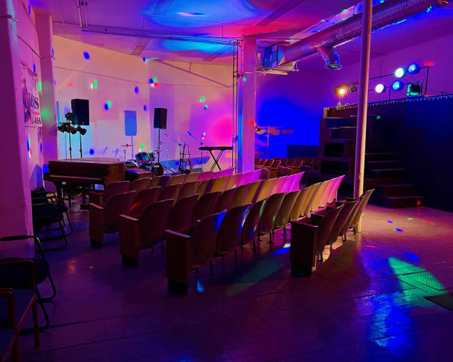 An empty performance space with an audience area of chairs, a stage with musical instruments, and colorful lights illuminating the room.