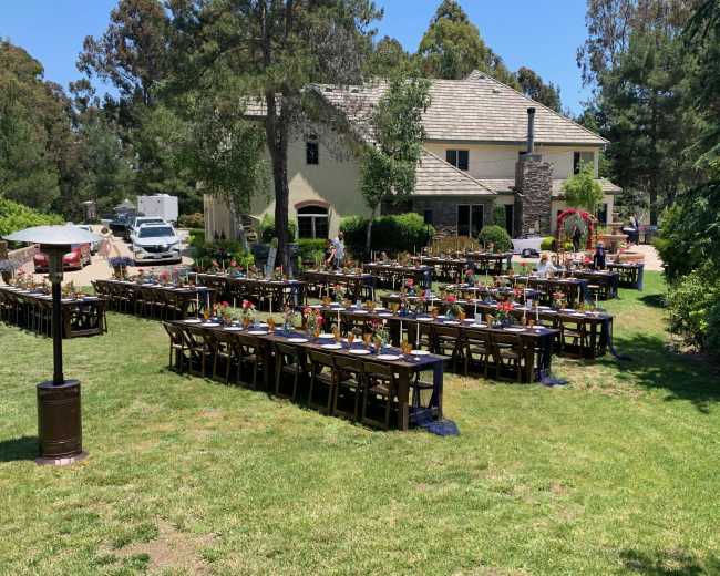 A large outdoor dining setup with several long tables is arranged on a grassy area in front of a spacious house, surrounded by trees.
