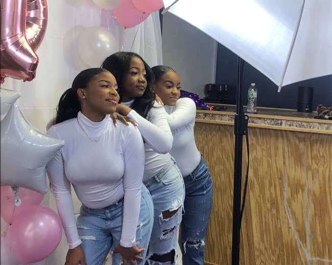 Three women pose together against a backdrop of balloons and decorations, dressed in white tops and distressed jeans, with a lighting umbrella nearby.