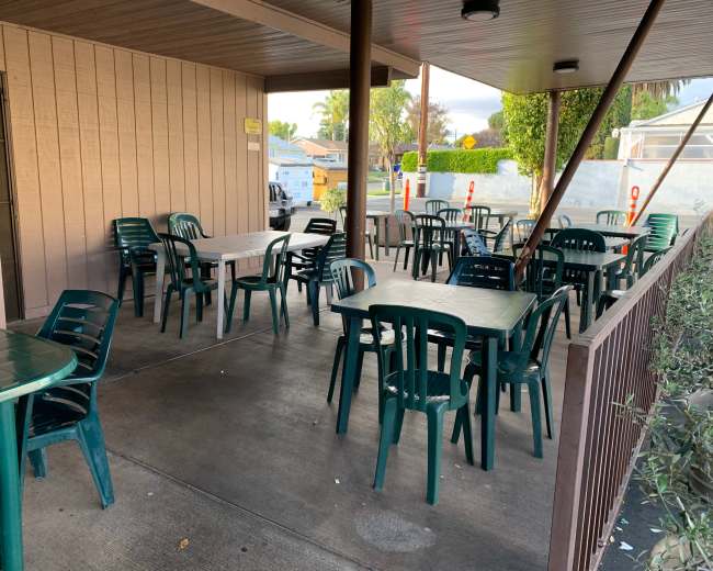 A covered outdoor seating area features several green plastic tables and chairs arranged on a paved surface.