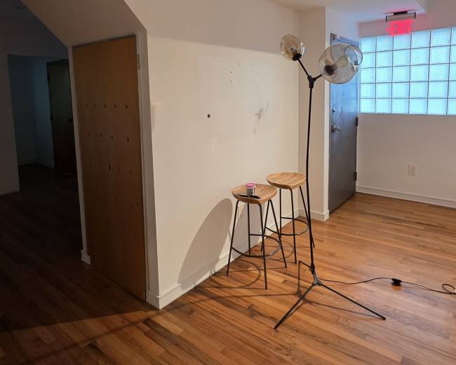 The image shows a sparsely furnished room with wooden flooring, featuring a tripod lamp and two stools near a white wall.