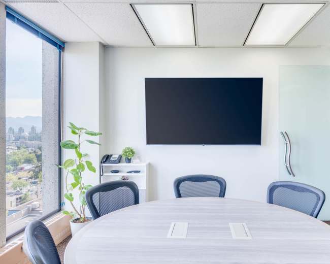 A modern conference room features a round table, several chairs, a wall-mounted television, and a large window with a city view.